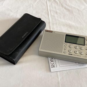 May include: A Roberts radio in grey, featuring a digital display and control buttons, alongside a black leather case. The radio displays the BBC World Service logo. A manual is also visible.