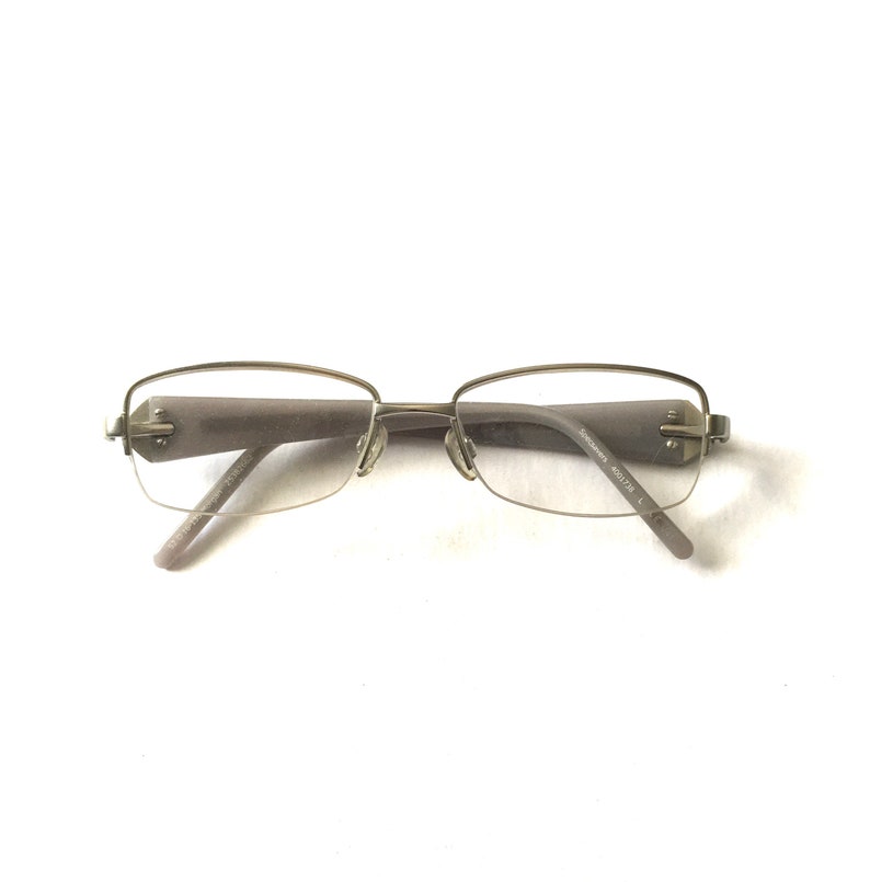 May include: Silver-toned rimless eyeglasses with a rectangular frame. The glasses have a subtle grey tint to the lenses.