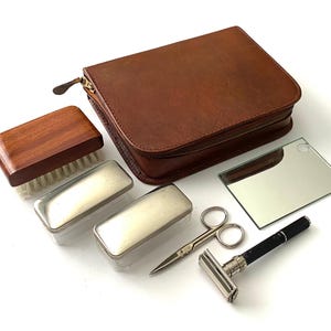 Puede incluir: Un neceser de cuero marrón con cierre de cremallera. El neceser contiene un cepillo de madera, dos recipientes de metal, un espejo pequeño, unas tijeras y una navaja de afeitar.