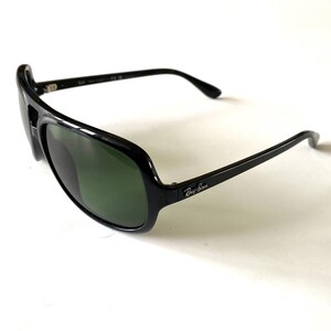 May include: Black sunglasses with green lenses. The sunglasses have a thick, glossy black frame and the Ray-Ban logo on the temple. The lenses are a dark green color, and the overall design is sporty and modern.