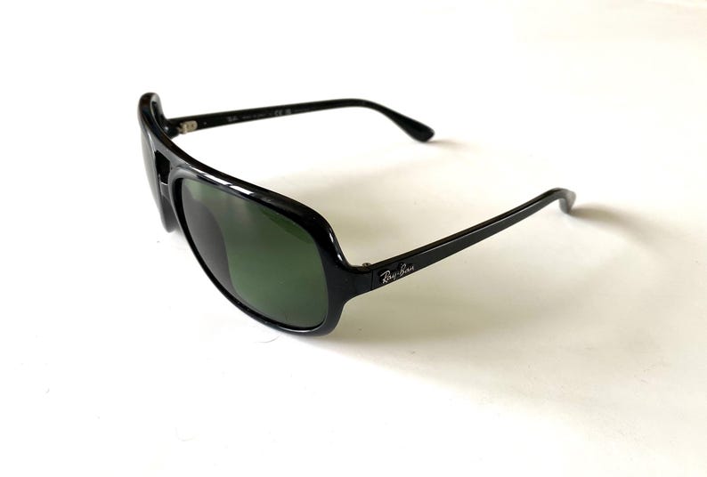 May include: Black sunglasses with green lenses. The sunglasses have a thick, glossy black frame and the Ray-Ban logo on the temple. The lenses are a dark green color, and the overall design is sporty and modern.