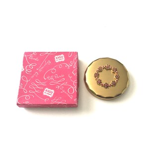 May include: A gold compact mirror with a pink flower design. The mirror is in its original box, which is pink with white lettering that says "Kigu of London" and "Pill Boxes".