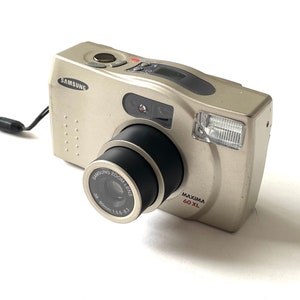 May include: A silver Samsung Maxima 60 XL point-and-shoot camera with a 38-60mm zoom lens. The camera has a black lens ring and a black strap.