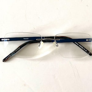 May include: Rimless eyeglasses with a navy blue top bar and temples. The frame is made of titanium. The lenses are clear and rectangular. The text "Titanium" is visible on the top bar.
