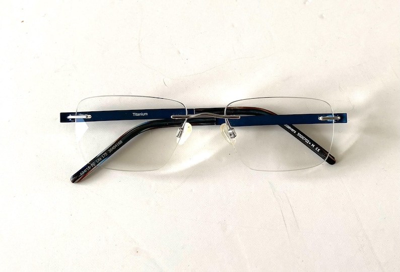 May include: Rimless eyeglasses with a navy blue top bar and temples. The frame is made of titanium. The lenses are clear and rectangular. The text "Titanium" is visible on the top bar.