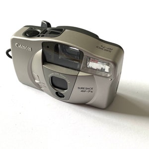 May include: A silver Canon Sure Shot AF-75 point-and-shoot camera with a 35mm lens. The camera has a built-in flash and a zoom lens.