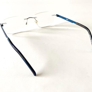 May include: Rimless eyeglasses with clear lenses and blue temples. The glasses have a silver bridge and hinges. The temples transition from blue to black. The glasses are designed for vision correction.