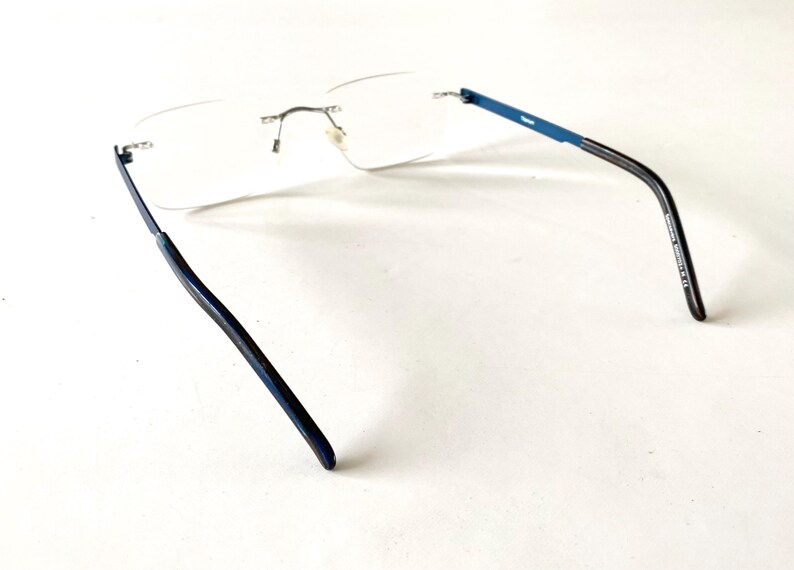 May include: Rimless eyeglasses with clear lenses and blue temples. The glasses have a silver bridge and hinges. The temples transition from blue to black. The glasses are designed for vision correction.