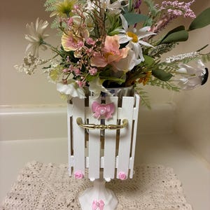 May include: A white picket fence-style vase with a gold handle and pink bow accents, filled with an assortment of colorful artificial flowers. The arrangement includes daisies, lavender, and other blooms, creating a cheerful, decorative display.
