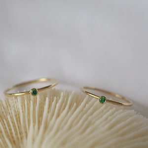 Può includere: Due delicati anelli d'oro con una singola pietra preziosa verde su ciascuno. Gli anelli sono esposti su una superficie bianca di tipo corallo.