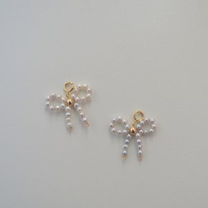 May include: Two gold charm pendants in the shape of bows made of white pearls. Each bow has a gold bead in the center.