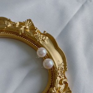May include: A pair of pearl stud earrings on a gold picture frame. The pearls are white and round.
