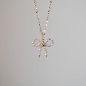 May include: A gold chain necklace with a pearl bow pendant. The bow is made of small white pearls and is tied in a knot at the center.