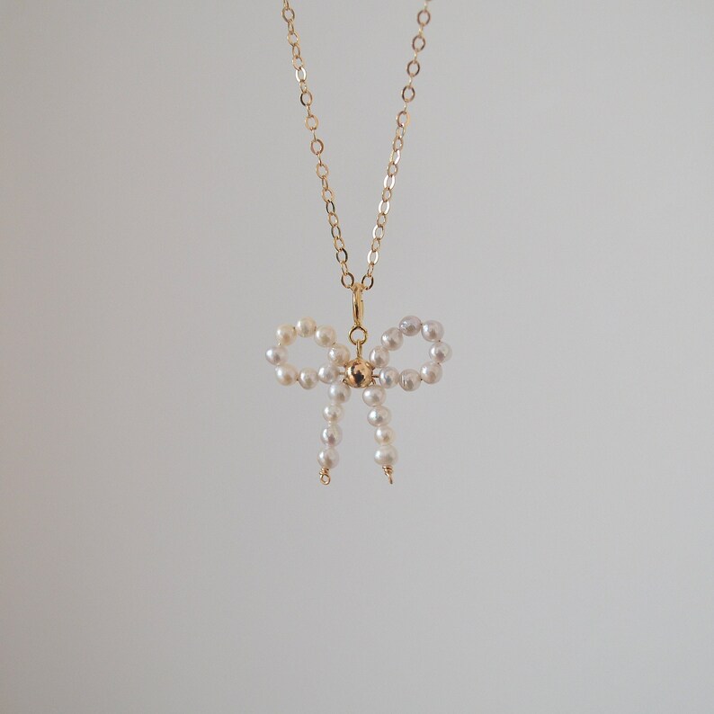 May include: A gold chain necklace with a pearl bow pendant. The bow is made of small white pearls and is tied in a knot at the center.