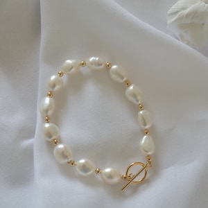 May include: A white pearl bracelet with a gold toggle clasp. The pearls are irregular in shape and have a smooth, glossy finish.