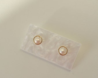 May include: A pair of pearl stud earrings. Each earring features a small, round, pale pink pearl set within a delicate gold-colored circular frame. The earrings are displayed on a rectangular, white and gray marbled surface.