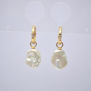 Może przedstawiać: Para złotych kolczyków koła z małymi, przezroczystymi kamieniami osadzonymi w kółkach. Każde kółko ma wiszącą perłę barokową.