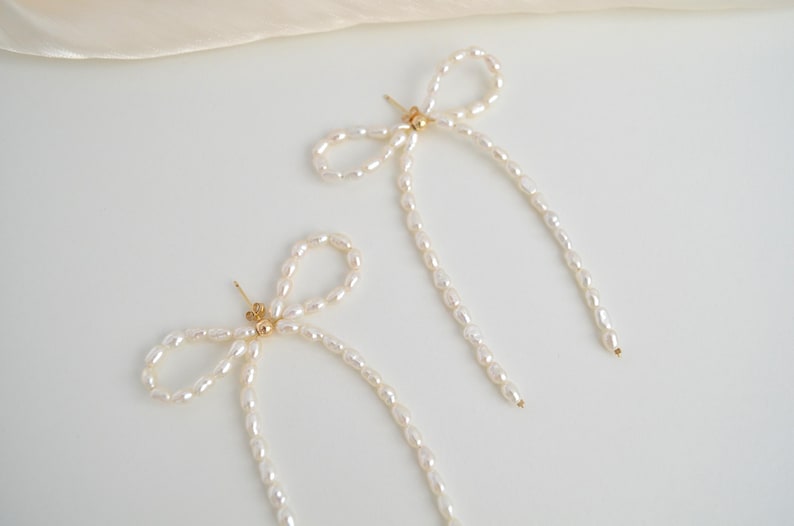 Pode incluir: Um par de brincos longos de p&eacute;rolas com um design de la&ccedil;o. Os brincos s&atilde;o feitos de pequenas p&eacute;rolas brancas enfiadas para formar um la&ccedil;o. O la&ccedil;o est&aacute; preso a uma corrente longa e fina de p&eacute;rolas que pende da orelha.