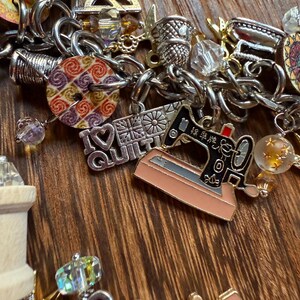 May include: A silver charm bracelet featuring a variety of charms, including a sewing machine, a spool of thread, a thimble, and a charm that reads "I Love Quilt".