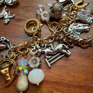 May include: A collection of equestrian-themed charms and beads in gold and silver tones. Charms include horses, horseshoes, and bits. The beads are white, clear, and iridescent. The charms are connected by gold-colored chains.