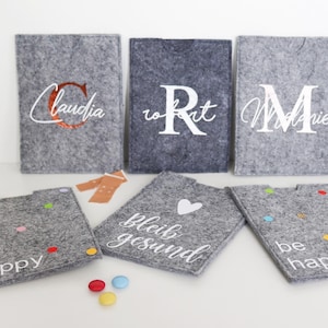 Könnte beinhalten: Sechs graue Filztaschen mit verschiedenen Stickereien. Die Taschen sind mit weißen Stickereien versehen, mit den Namen "Claudia", "Robert", "Mollie", "be happy", "be happy" und "bleib gesund". Eine Tasche hat ein weiß gesticktes Herz.