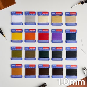 Könnte beinhalten: Zwanzig Spulen Ritza 25 Julius Koch gewachster Faden in einem Raster angeordnet. Jede Spule hat einen blauen Papphalter mit weißem Text und zeigt eine Vielzahl von Farben, darunter Weiß, Grau, Beige, Gelb, Orange, Rot, Lila, Blau, Grün, Braun und Schwarz. Der Fadendurchmesser beträgt 1,0 mm.