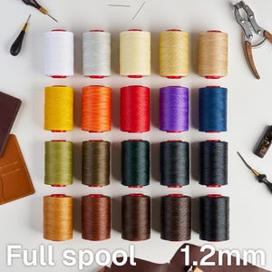 Op de afbeelding: Een collectie van twintig klossen garen van 1,2 mm gerangschikt in een raster, met een verscheidenheid aan kleuren waaronder wit, geel, oranje, rood, paars, blauw, groen, bruin en zwart. Elke klos heeft rode uiteinden en is omringd door ledergereedschap.