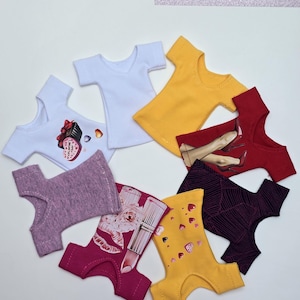 Può includere: Una collezione di vestiti per bambole, tra cui magliette bianche, gialle e rosse e vari pantaloni fantasia. Alcuni pantaloni presentano rossetto, cuori e tacchi alti. L'immagine include il testo "DzastPrince".