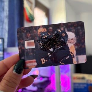 May include: A rectangular card featuring a hockey team image, with players in black and yellow uniforms. The card has a white rectangle on the left side. The image is held by a hand with a dark green nail polish.