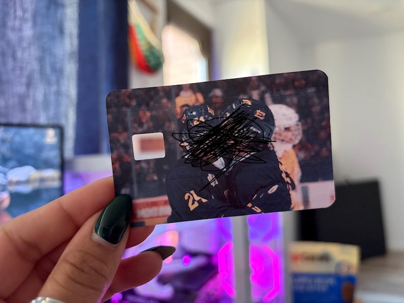 May include: A rectangular card featuring a hockey team image, with players in black and yellow uniforms. The card has a white rectangle on the left side. The image is held by a hand with a dark green nail polish.