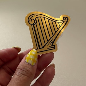 Könnte beinhalten: Ein goldfarbener Harfenaufkleber mit schwarzen Umrissen. Der Aufkleber hat ein klassisches Harfendesign mit einem geschwungenen Oberteil und vertikalen Saiten. Der Aufkleber wird von einer Hand mit gelb-weiß kariertem Nagellack gehalten.