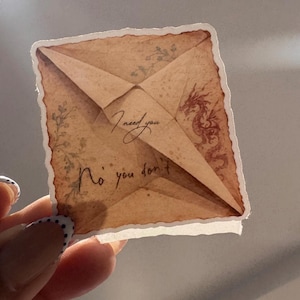 May include: A square sticker with a vintage paper aesthetic. The sticker features a folded paper design with the text "I need you" and "No you don't". A dragon illustration and floral accents are also visible.