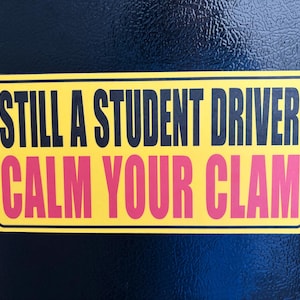Op de afbeelding: Een rechthoekige gele bumpersticker met een zwarte rand. De tekst luidt STILL A STUDENT DRIVER in zwarte hoofdletters op de bovenste regel en CALM YOUR CLAM in dikgedrukte roze hoofdletters op de onderste regel van de sticker.