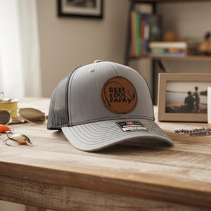 May include: Gray trucker hat with a black mesh back and a brown leather patch that reads "Reel Cool Papa." The hat is on a wooden table with fishing lures and sunglasses.