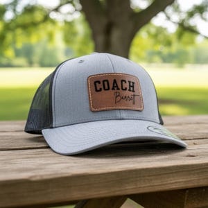 May include: A gray and black trucker hat with a brown leather patch. The patch has the word "COACH" above the name "Barrett" in a script font. The hat is sitting on a wooden picnic table.