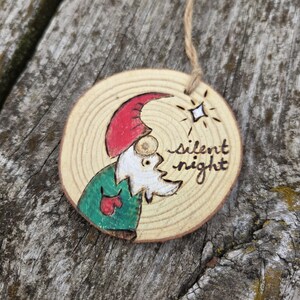 May include: Wooden Christmas ornament featuring a hand-painted gnome with a red hat and green coat. The ornament is round, made from a wood slice, and has the words "silent night" written on it. A small star is drawn above the text. A twine loop is attached for hanging.