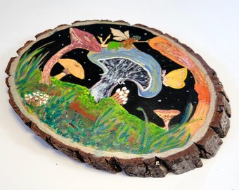 May include: A whimsical, hand-painted wooden slice featuring a fantasy scene. The artwork depicts colorful mushrooms, a fairy, and a butterfly against a starry black background. The wood has a natural, rough bark edge, adding a rustic touch.
