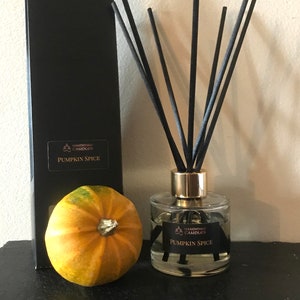May include: A clear glass bottle with a gold lid and black reeds, filled with a light yellow liquid. The bottle has a black label that reads "Handmade Candles Pumpkin Spice". A small orange pumpkin sits next to the bottle.