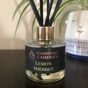 May include: A clear glass diffuser bottle with a gold-coloured metal collar and black reeds. The label reads "Handmaid Candles Lemon Sherbet" in gold and pink lettering. The bottle is on a dark surface.