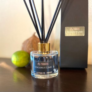 May include: A clear glass diffuser bottle with black reeds and a gold-coloured collar. The label reads "Handmade Candles Coconut Lime." A lime and a coconut are in the background. A black box with a gold label that reads "Coconut Lime" is to the right.