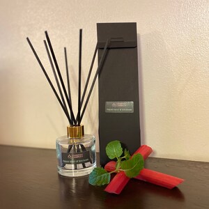 May include: A glass diffuser bottle with black reeds and a gold collar, labelled "Fresh Mint & Rhubarb." A black box and fresh rhubarb with mint leaves are next to the bottle. The product is from "Handmade Candles."
