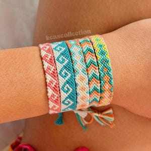 May include: A collection of colorful woven friendship bracelets. The bracelets feature geometric patterns in shades of pink, turquoise, orange, and yellow. Each bracelet is approximately 0.5 inches wide. The bracelets are tied with tassels.