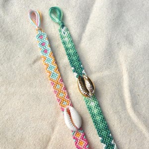 May include: Two handmade friendship bracelets with colorful woven patterns. One bracelet features a white seashell, the other a gold seashell. The bracelets are in shades of pink, yellow, blue, and green.