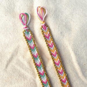May include: Two handmade friendship bracelets. Each bracelet features a chevron pattern in pink, white, green, and yellow, outlined with gold thread. The ends are looped and knotted with matching colors, creating a decorative finish.