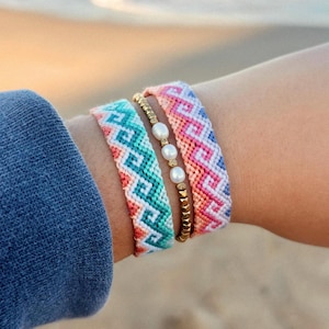 May include: A collection of colorful woven friendship bracelets. The bracelets feature geometric patterns in shades of pink, blue, green, and white. One bracelet has gold beads and pearls.