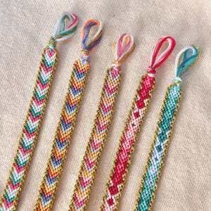 May include: Five colorful friendship bracelets with chevron patterns. Each bracelet features a different color scheme, including teal, pink, orange, and blue, with gold-colored edging. The bracelets are knotted at the top with matching thread.