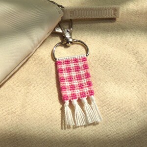 May include: A pink and white woven keychain with a gingham pattern and three white tassels. The keychain is attached to a silver metal clasp and a beige fabric. The woven design is a repeating pattern of pink and white squares.