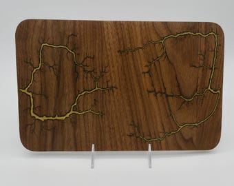 May include: Rectangular wooden serving board with rounded corners. The dark brown wood features a unique design of yellow lightning-like patterns. The board is displayed on a clear stand, showcasing its decorative appeal.
