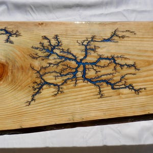 May include: A rectangular wooden slab with a natural edge. The light-colored wood features a prominent knot and is decorated with blue fractal patterns resembling lightning. The wood grain is visible, and the surface has a glossy finish.