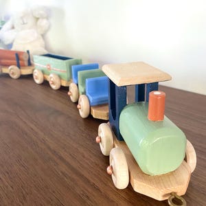Könnte beinhalten: Ein hölzernes Spielzeugeisenbahn-Set mit einer hellgrünen Lokomotive mit orangefarbenem Schornstein, einem blauen Personenwagen, einem grünen Güterwagen und einem Flachwagen mit orangefarbenen Holzstämmen. Die Räder aus Naturholz haben kupferfarbene Achsen.